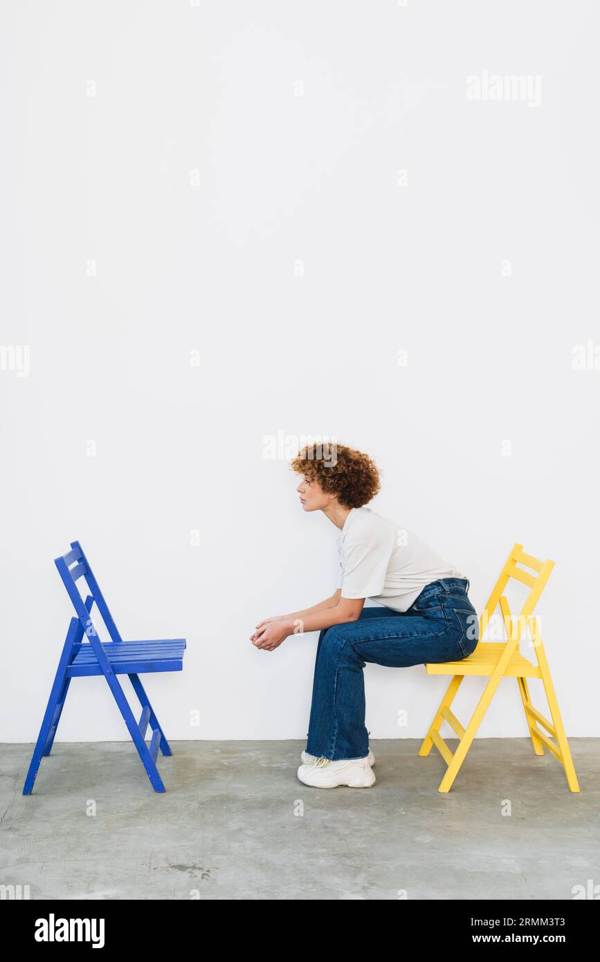 Femme de gingembre d'âge moyen posant avec deux chaises isolées sur le mur blanc Banque D'Images