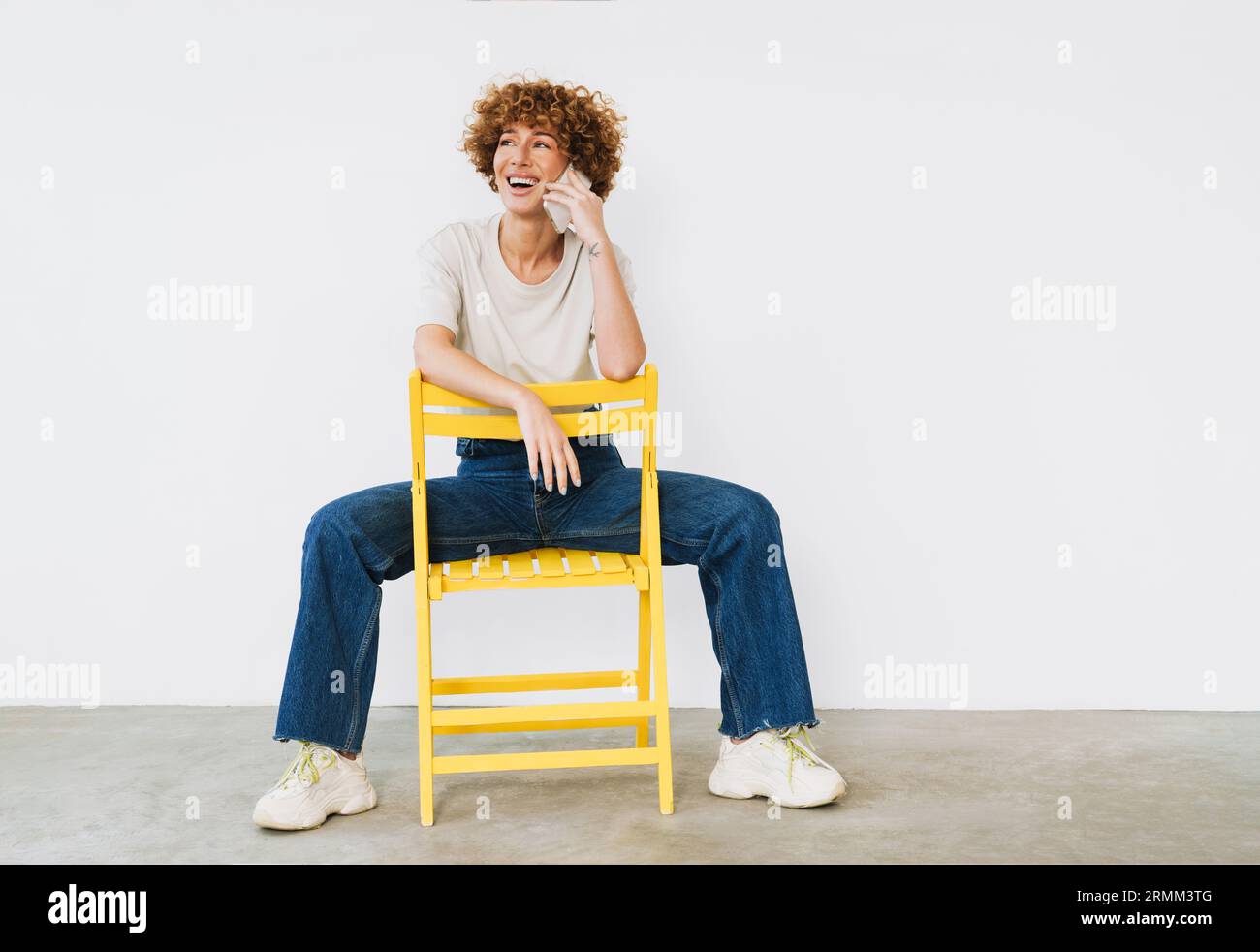 Joyeuse femme mature assise sur une chaise jaune et parlant sur un téléphone portable isolé sur un mur blanc Banque D'Images
