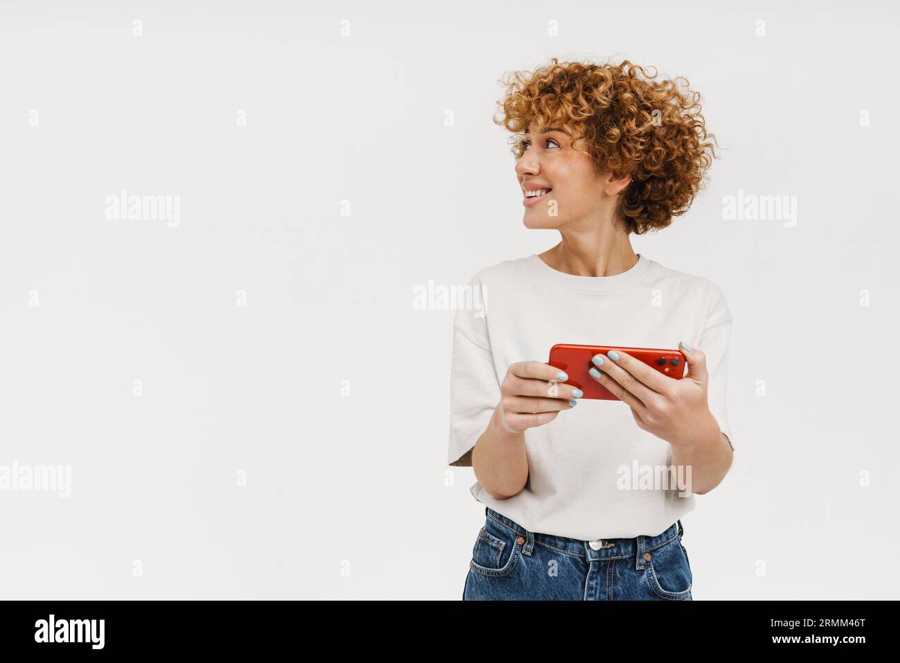 Femme bouclé de gingembre portant un t-shirt souriant et utilisant un téléphone portable isolé sur fond blanc Banque D'Images