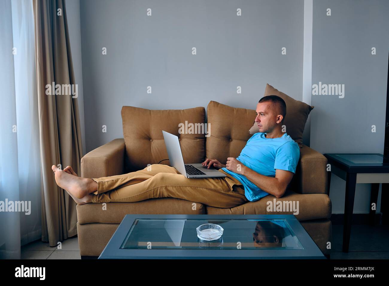 Jeune homme caucasien adulte travaille sur un ordinateur portable allongé sur un canapé. Homme réfléchi surfant sur un filet couché sur le canapé Banque D'Images