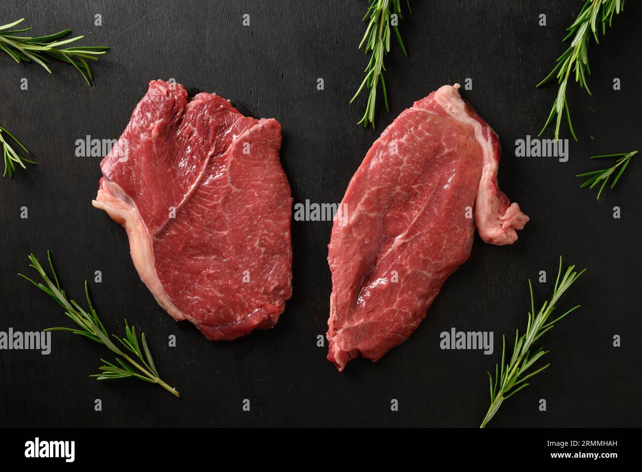 Cuisson du steak de ribeye marbré cru avec branche de romarin sur une planche à découper sur fond noir. Vue de dessus. Banque D'Images