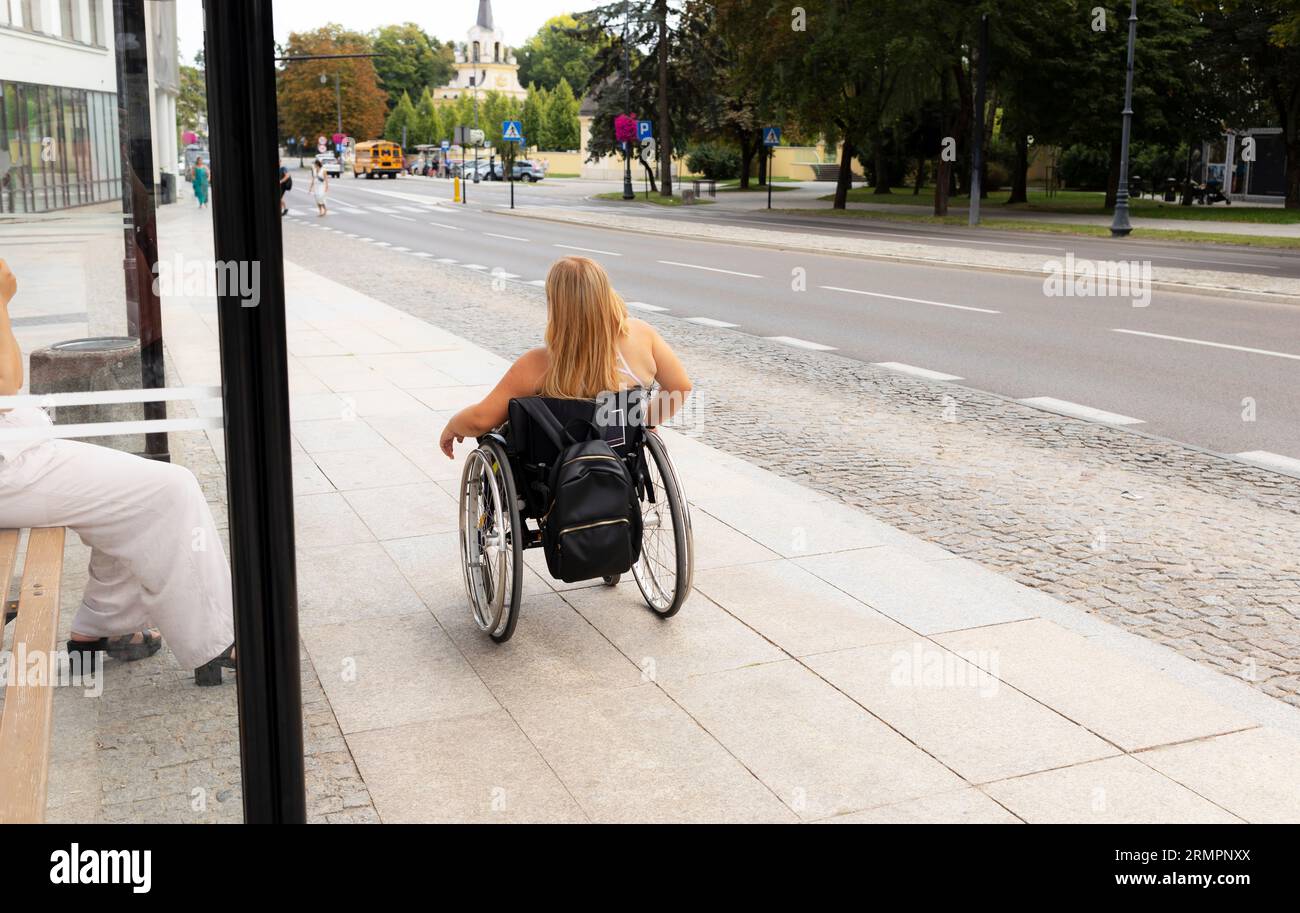 Vue arrière adulte avec petite taille sur les stands en fauteuil roulant sur l'arrêt de bus extérieur en attente des transports publics le jour de l'été. Femme adulte handicapée Banque D'Images