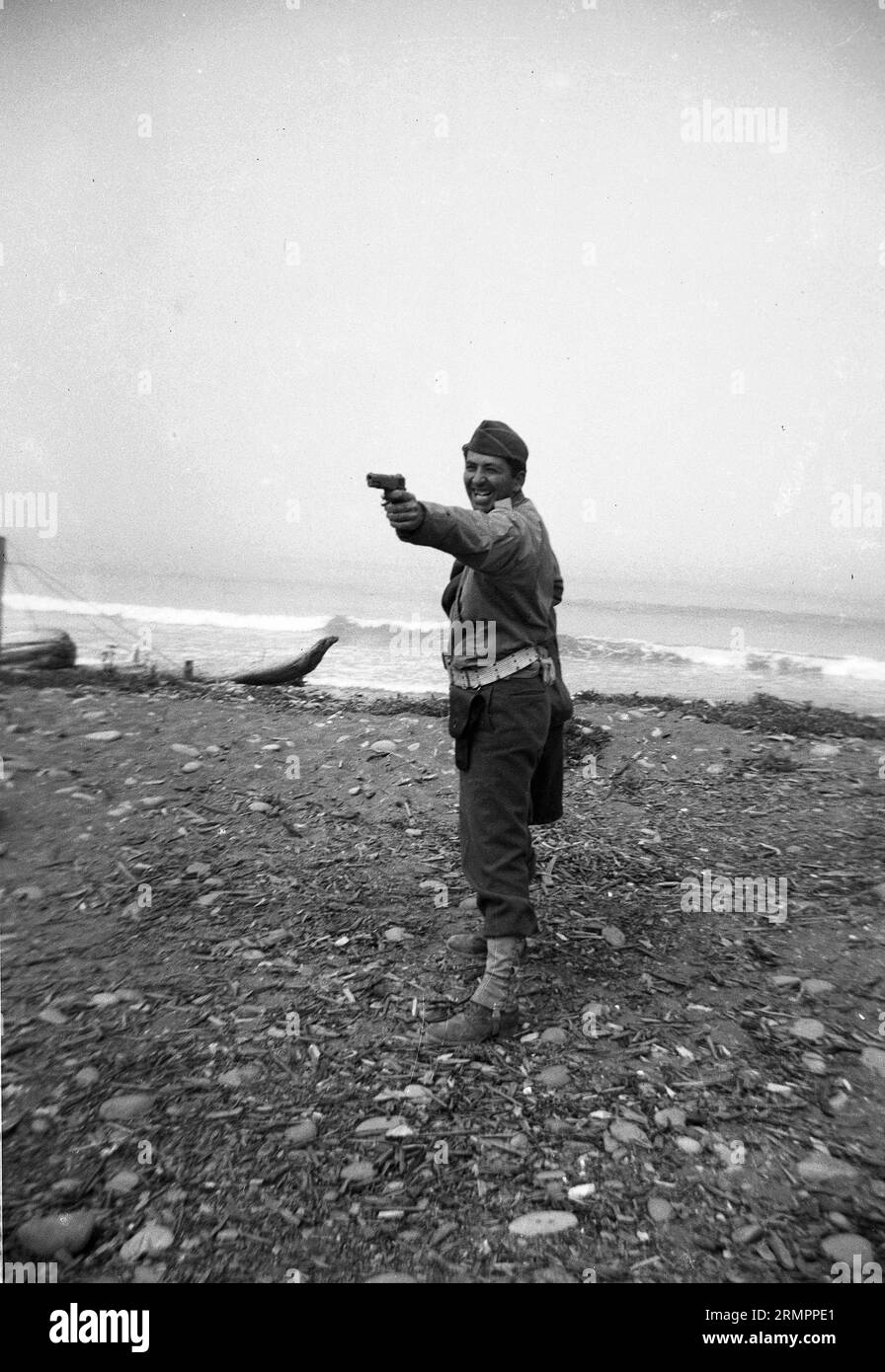 Solider posant et riant tout en pointant 1911 .45 pistolet près de l'océan. Les membres de la 114e division d’infanterie de l’armée des États-Unis s’entraînent pour combattre l’Allemagne en Europe pendant la Seconde Guerre mondiale. Banque D'Images