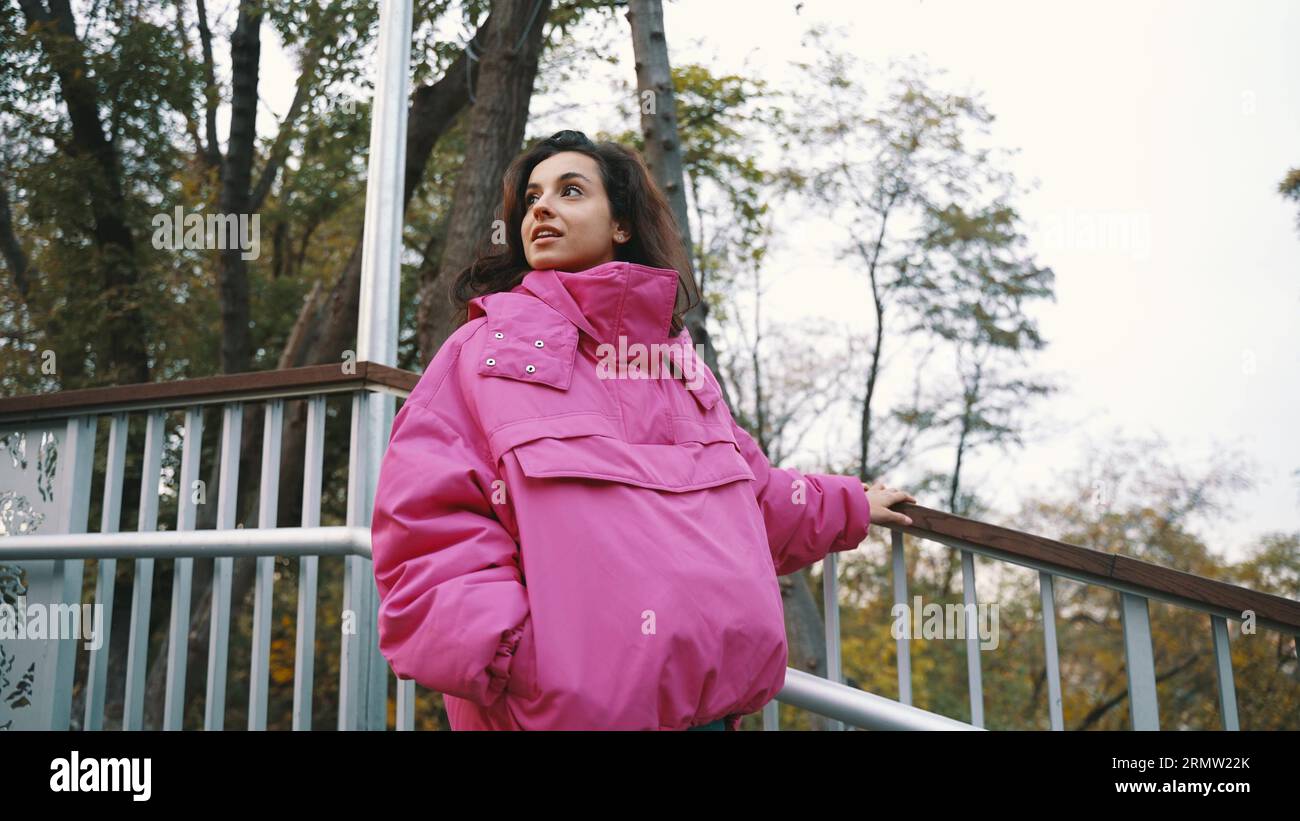 Jolie jeune femme descendant les escaliers tout en marchant dans le parc, se tenant à la rampe, portant une veste rose élégante. Style saisonnier, saison d'automne Banque D'Images