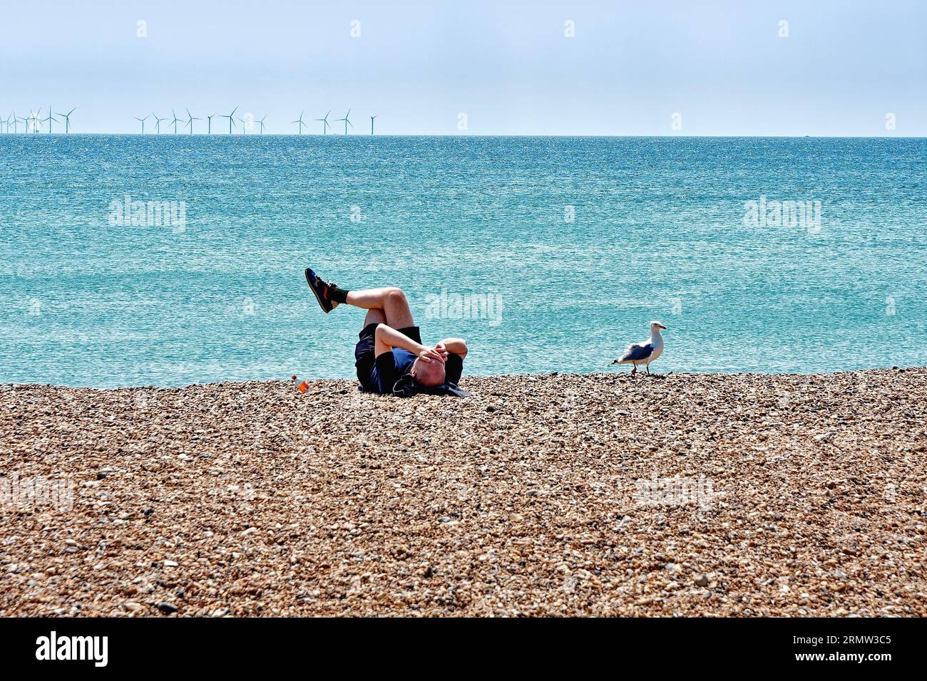 Un jeune homme allongé sur la plage de galets bronzer avec une goélette à hareng assise à côté de lui, avec un parc éolien à l'horizon, Worthing Sussex Angleterre Royaume-Uni Banque D'Images