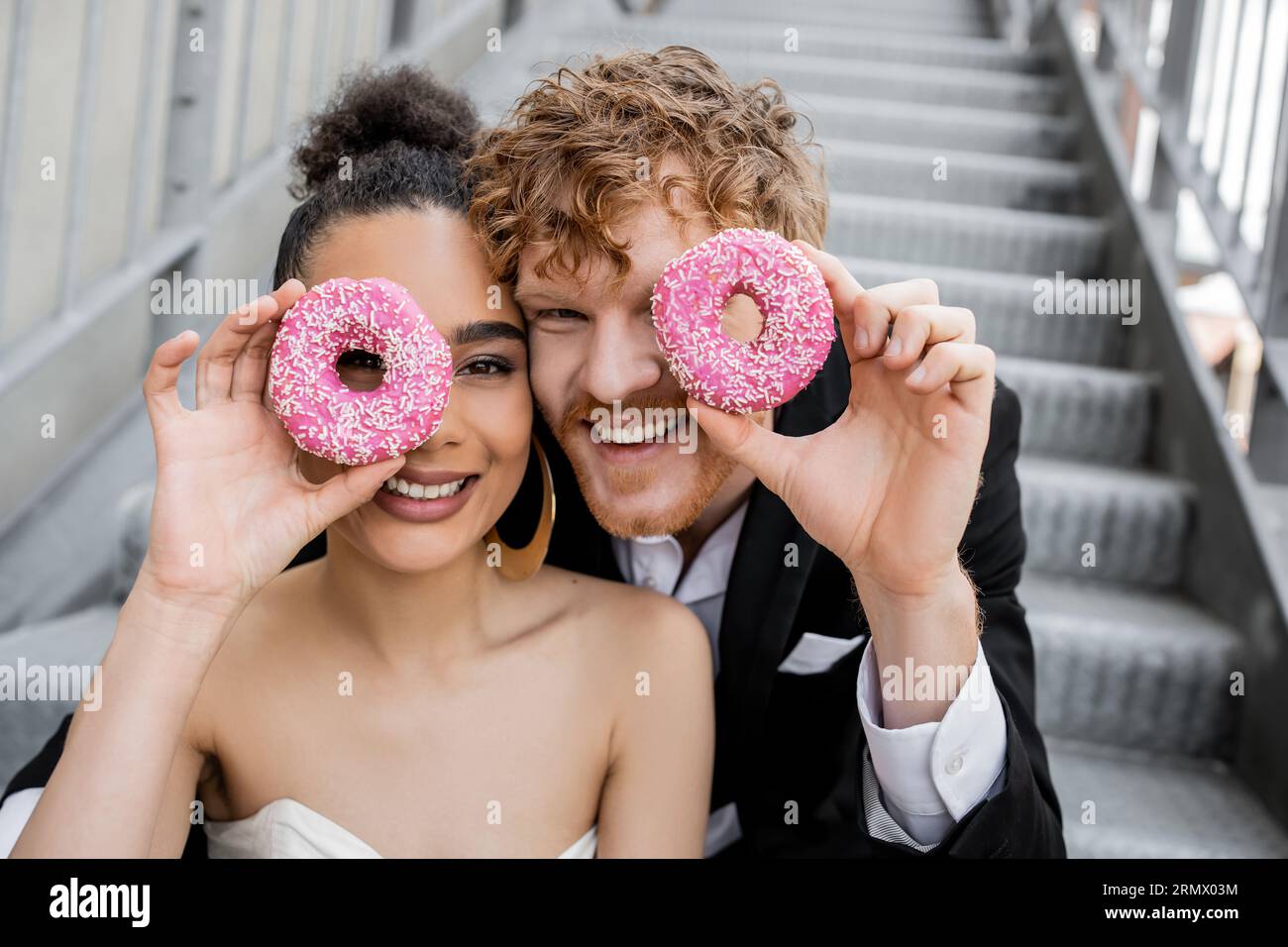 s'amuser, se marier en ville, excités jeunes mariés interraciaux obscurcissant le visage avec des beignets Banque D'Images