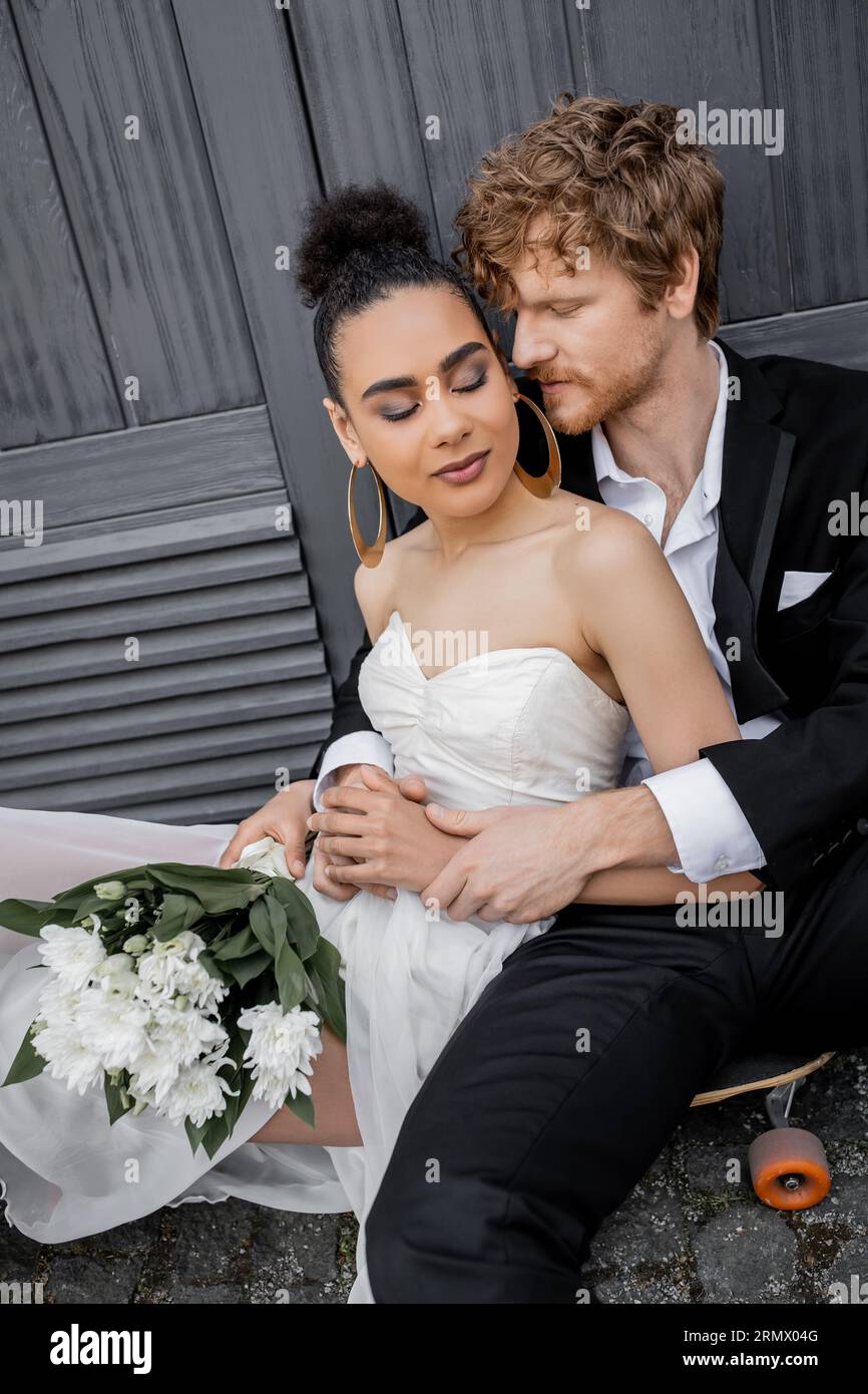 le marié redhead embrasse la mariée afro-américaine avec les yeux fermés et les fleurs, assis près du bâtiment Banque D'Images