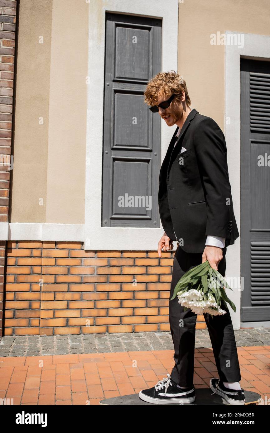 homme élégant en costume noir et lunettes de soleil marchant dans la rue de la ville, marié avec bouquet de mariage Banque D'Images