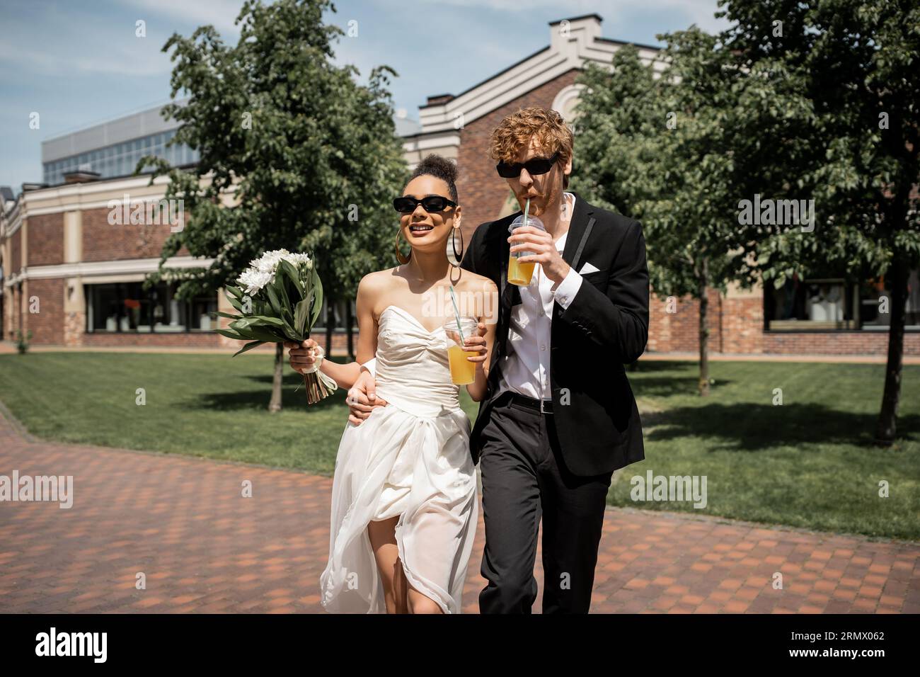 élégants jeunes mariés dans des lunettes de soleil marchant avec du jus d'orange et des fleurs dans la ville européenne Banque D'Images