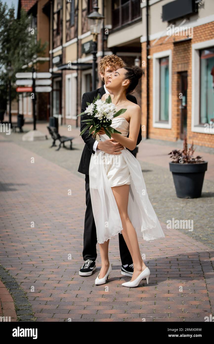 jeune homme rousse embrassant femme afro-américaine en robe de mariée, fleurs, joie, mariage en ville Banque D'Images