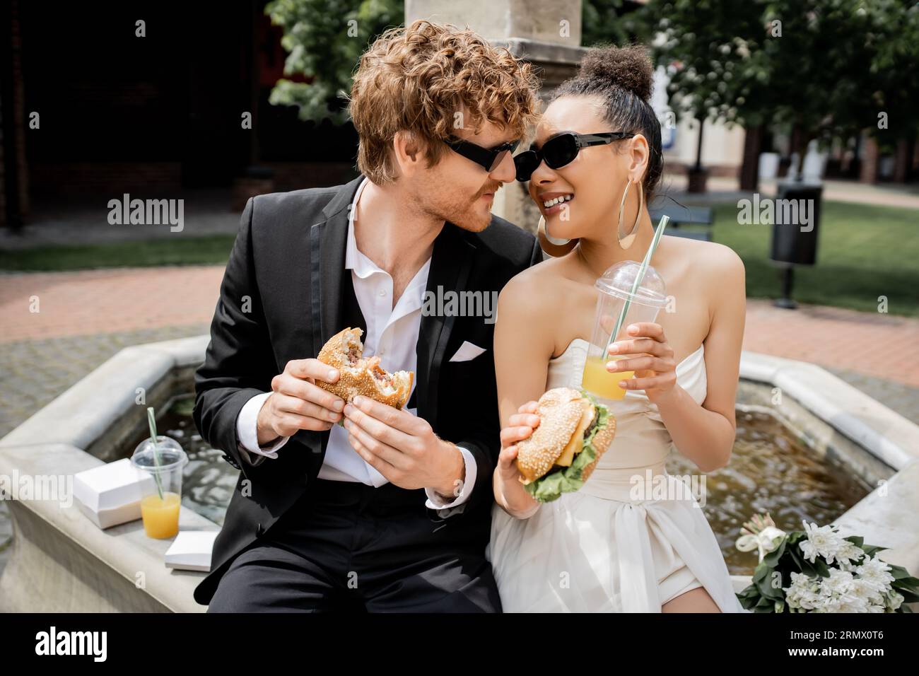 élégants jeunes mariés interraciaux en lunettes de soleil avec hamburgers et jus d'orange près de la fontaine de la ville Banque D'Images