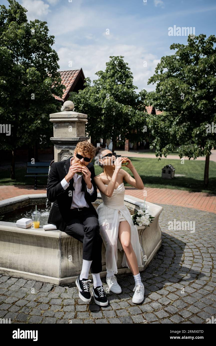 élégants jeunes mariés interraciaux dans des lunettes de soleil mangeant des hamburgers près de la fontaine dans la ville européenne Banque D'Images