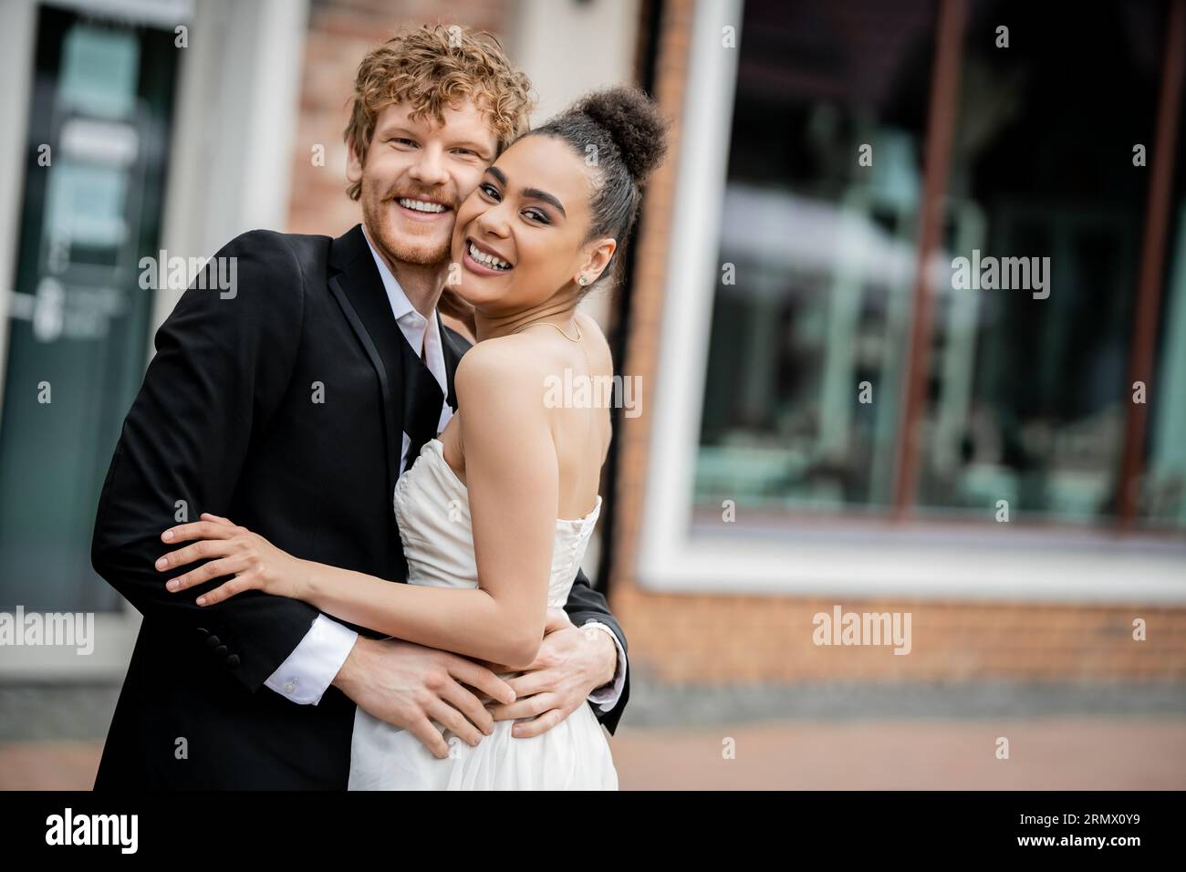joyeux et élégant jeunes mariés multiethniques étreignant et regardant la caméra dans la rue, mariage en plein air Banque D'Images