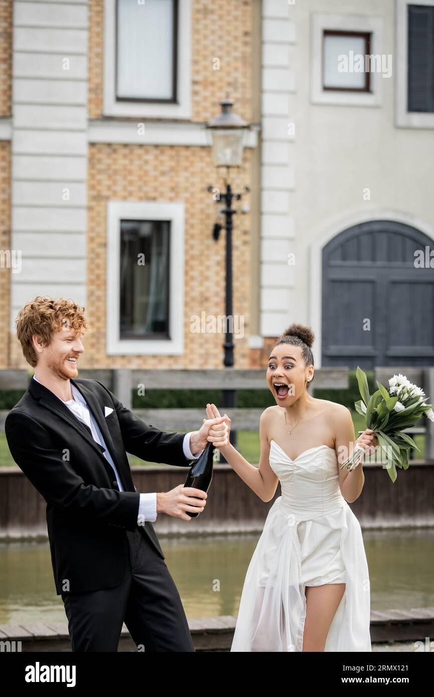 mariage en ville, marié rousse avec champagne versé de bouteille près de la mariée afro-américaine criante Banque D'Images