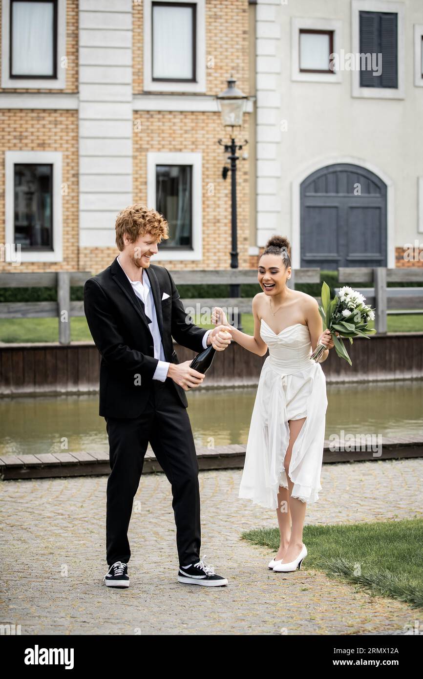 mariage en ville, marié ouvrant champagne près de mariée afro-américaine excitée avec des fleurs sur la rue Banque D'Images