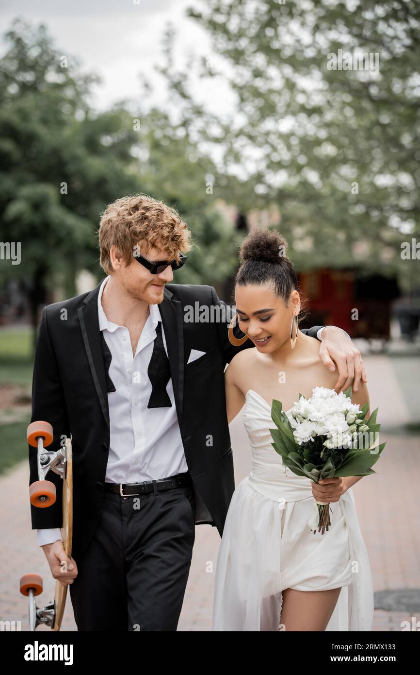 redhead groom dans des lunettes de soleil embrassant la mariée afro-américaine, marchant avec longboard et fleurs Banque D'Images