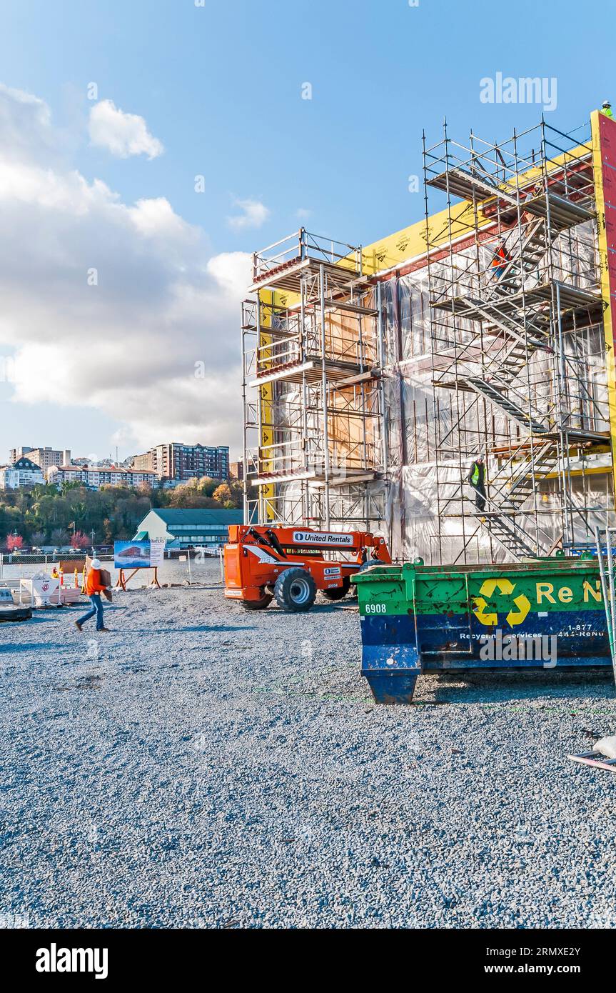 Un haut-ascenseur de location orange avec son bras étendu au toit d'un bâtiment sur un chantier de construction, tandis qu'un travailleur de la construction dans un casque de dur marche sur le site. Banque D'Images