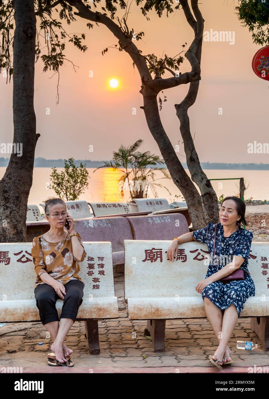 Pakse, Sud Laos-février 12 2023 : assis sur des bancs en métal inscrits chinois, près de la rivière, les dames se détendent et socialisent, à l'approche du crépuscule, en Banque D'Images
