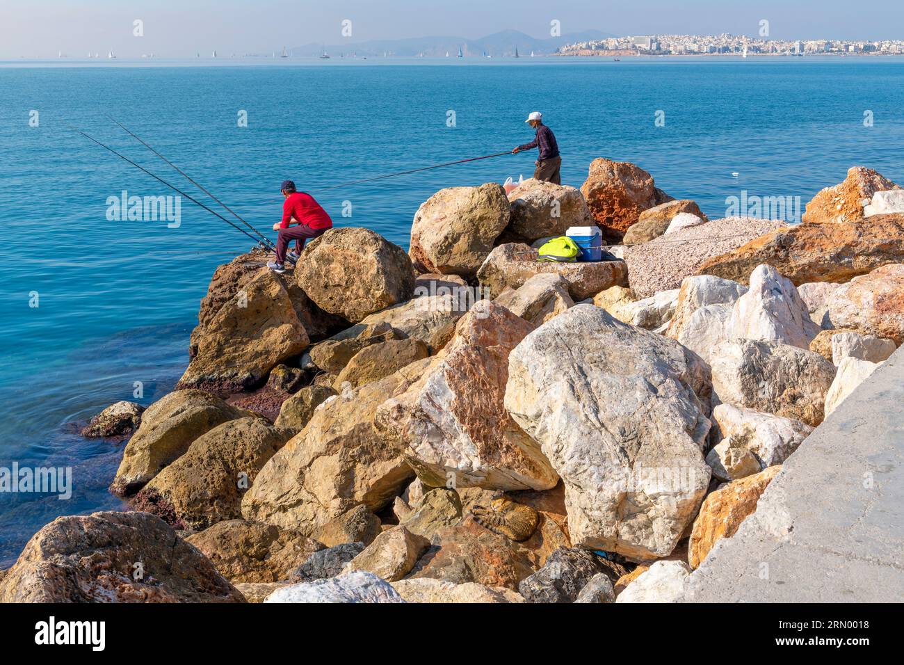 Deux hommes pêchent au large des rochers de bord de mer et un chat se fond dans les rochers en contrebas dans la région de la Riviera athénienne, Paleo Faliro, près d'Athènes, Grèce. Banque D'Images