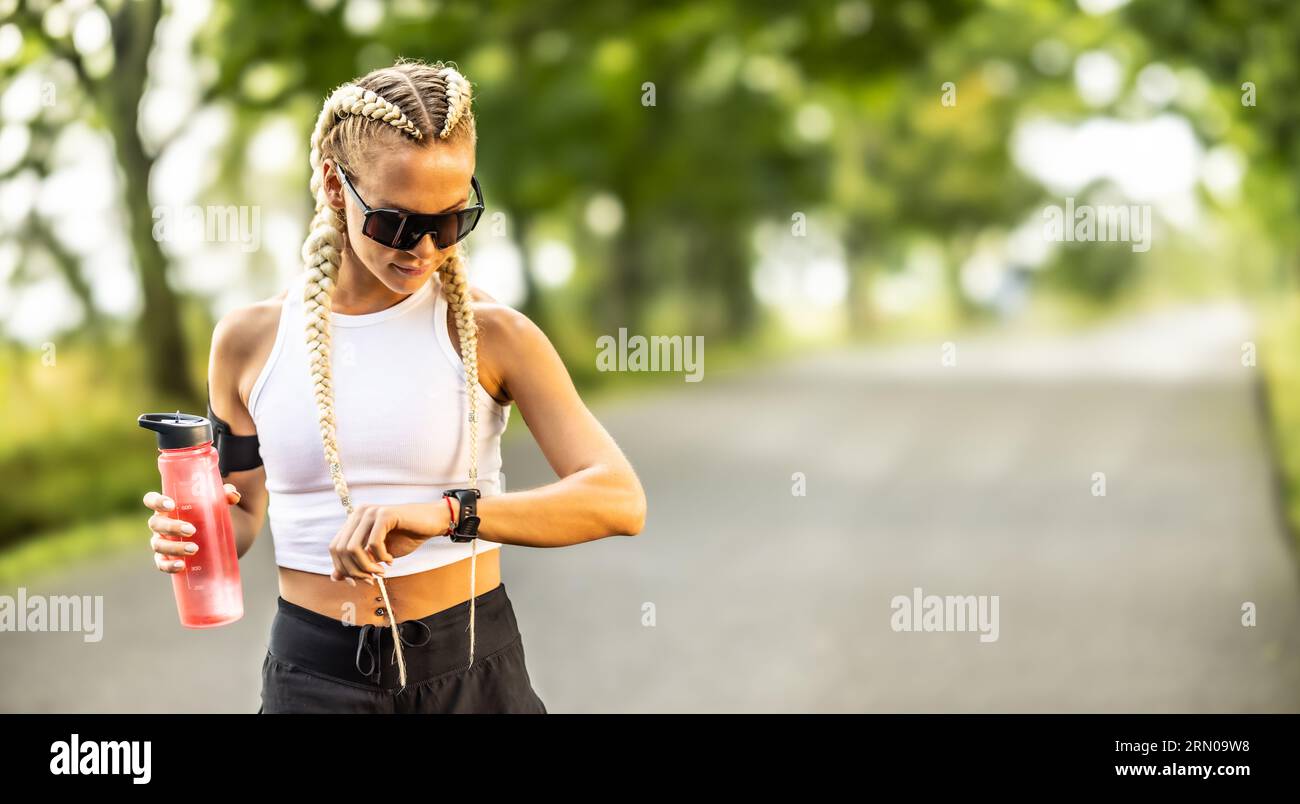 Belle athlète féminine vérifiant sa fréquence cardiaque sur la montre intelligente, tenant la bouteille d'eau pour l'hydratation avant le marathon. Banque D'Images