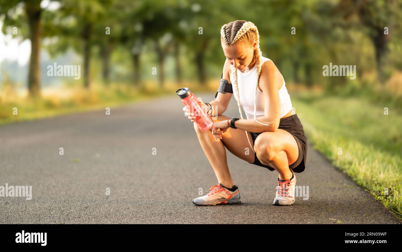 Belle athlète féminine vérifiant sa fréquence cardiaque sur la montre intelligente, tenant la bouteille d'eau pour l'hydratation, tout en se reposant. Banque D'Images