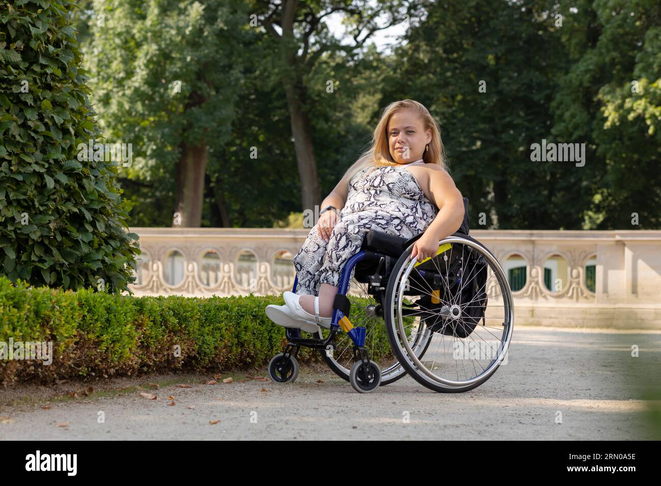 Full Length Young Pretty Woman avec petite stature sur fauteuil roulant regardant la caméra dans le parc public le jour d'été. Sourire Pretty Female adulte avec Banque D'Images