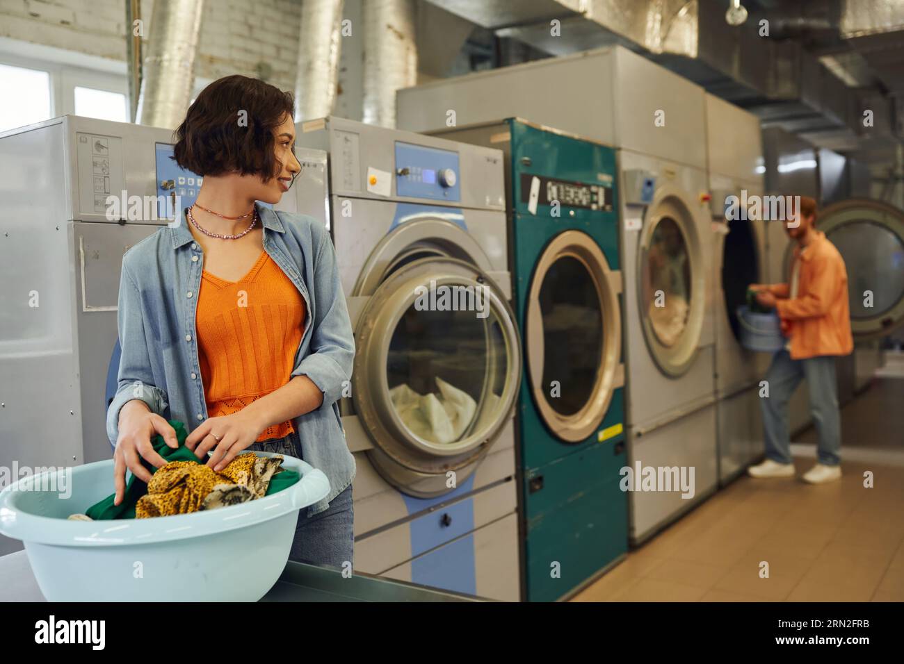 jeune femme asiatique souriante regardant petit ami près des vêtements dans le bassin dans le linge flou de pièce Banque D'Images