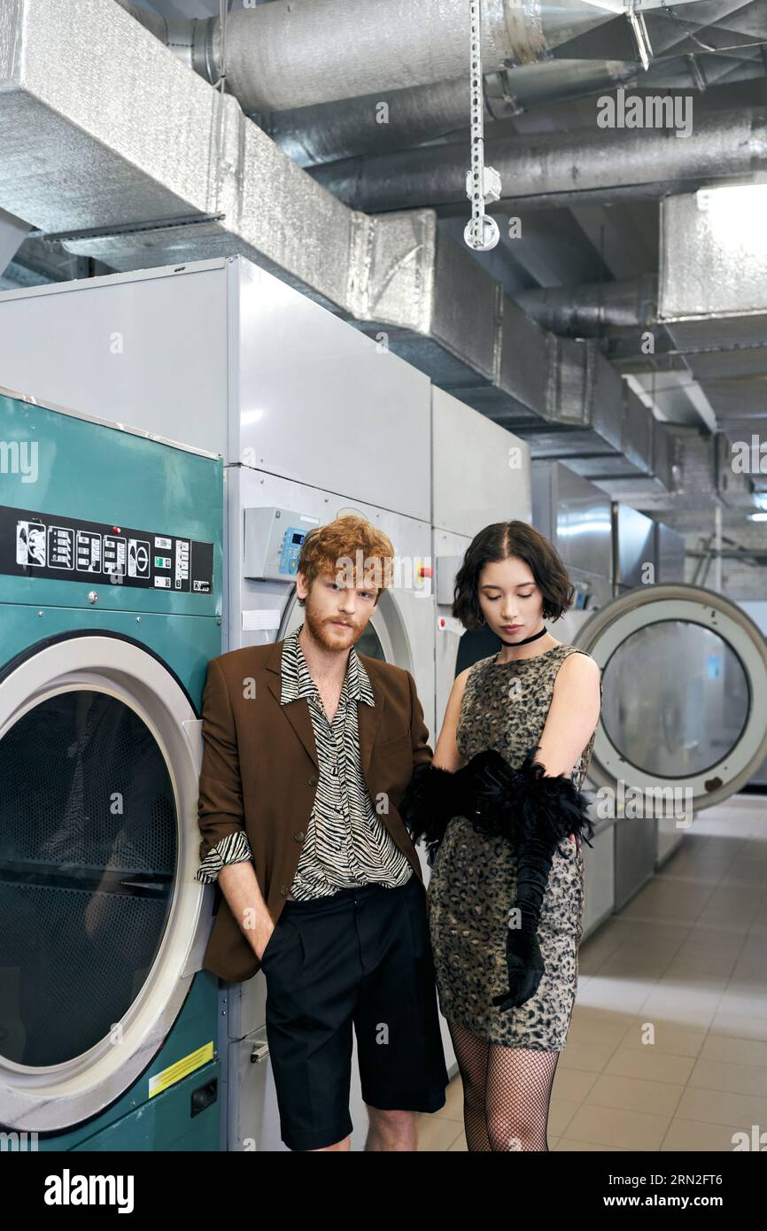 jeune couple multiethnique à la mode debout près des machines à laver dans la blanchisserie publique Banque D'Images