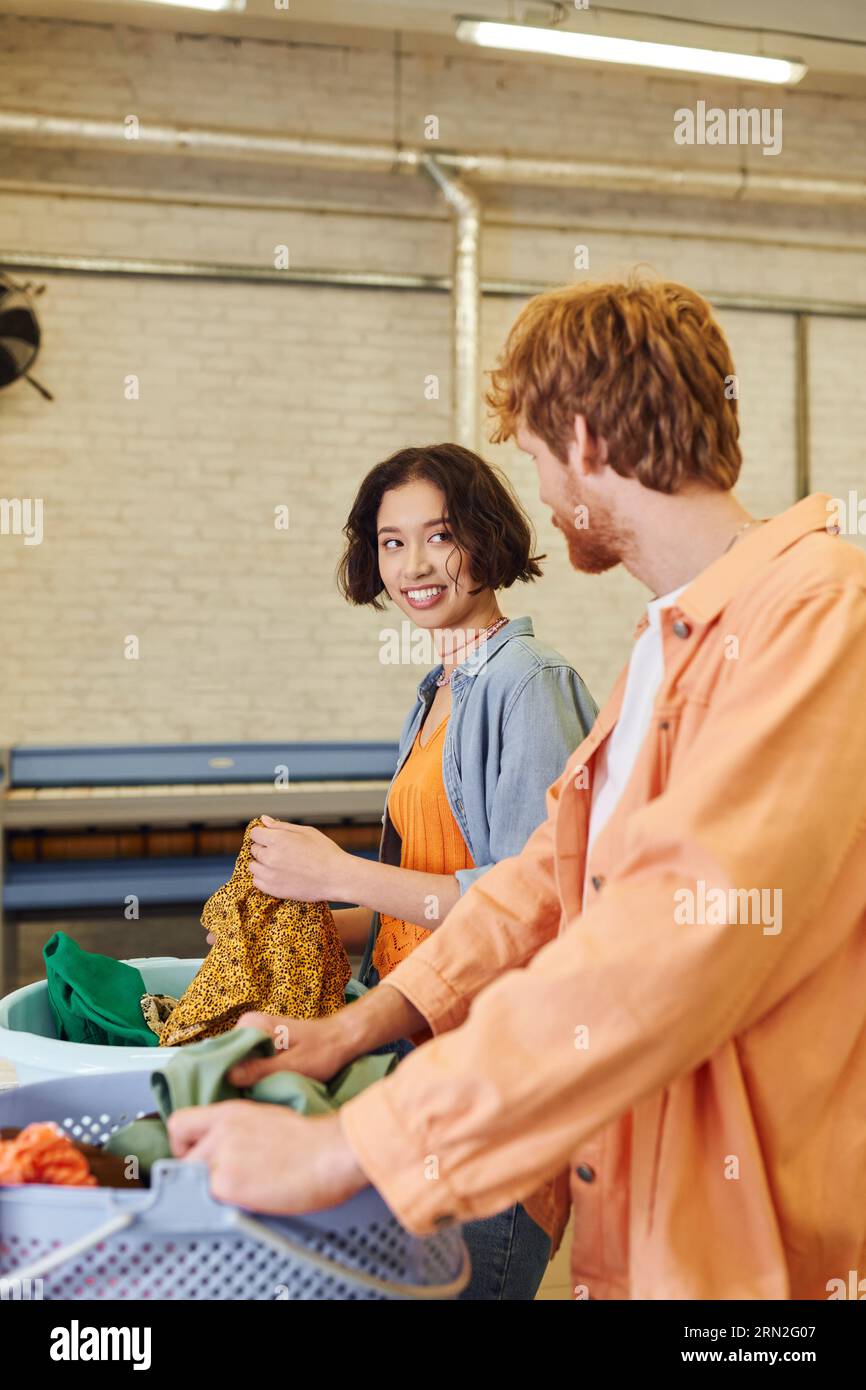 joyeuse jeune femme asiatique parlant à son petit ami près des vêtements et des paniers dans le linge à pièces Banque D'Images