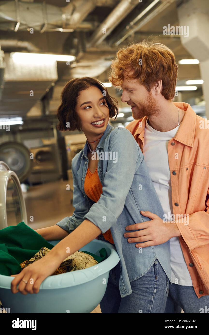 redhead homme embrassant joyeuse petite amie asiatique avec des vêtements dans le bassin dans la blanchisserie publique Banque D'Images