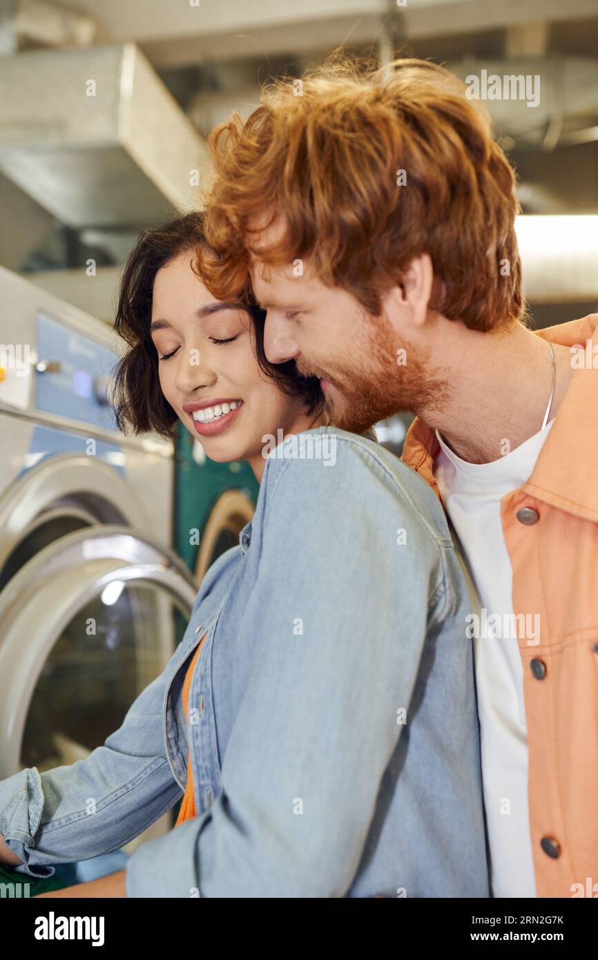 jeune rousse embrassant une joyeuse petite amie asiatique dans une blanchisserie publique floue Banque D'Images
