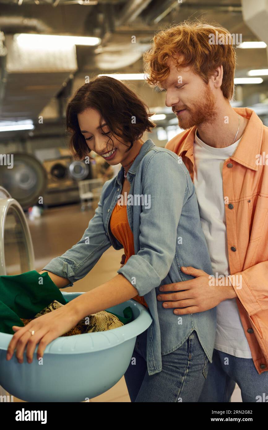 redhead homme embrassant joyeuse petite amie asiatique avec des vêtements et un panier dans le linge de pièce Banque D'Images