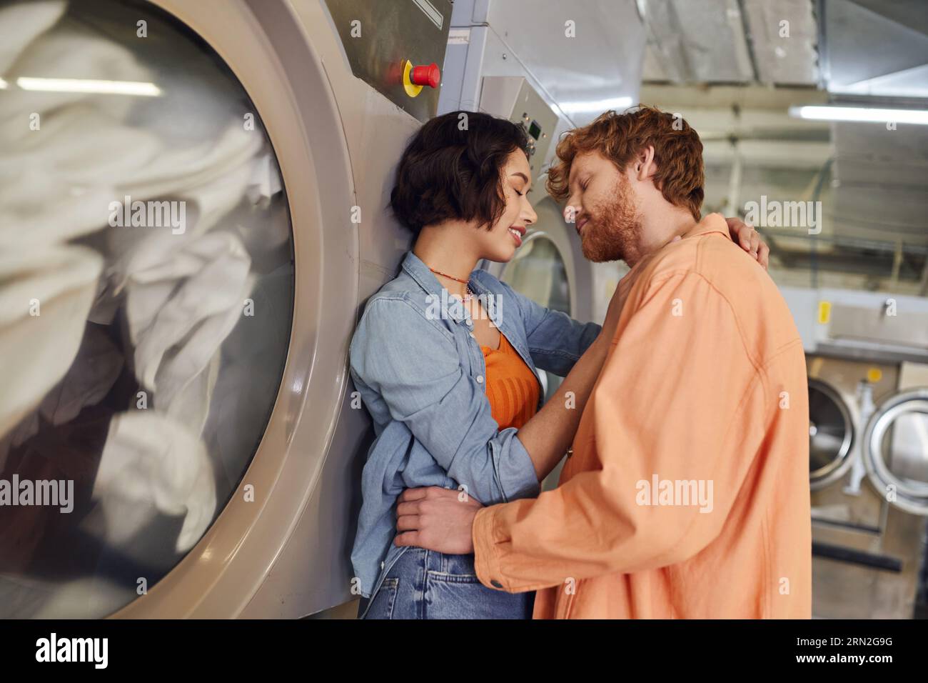 femme asiatique souriante embrassant petit ami rousse près de la machine à laver dans la blanchisserie publique Banque D'Images