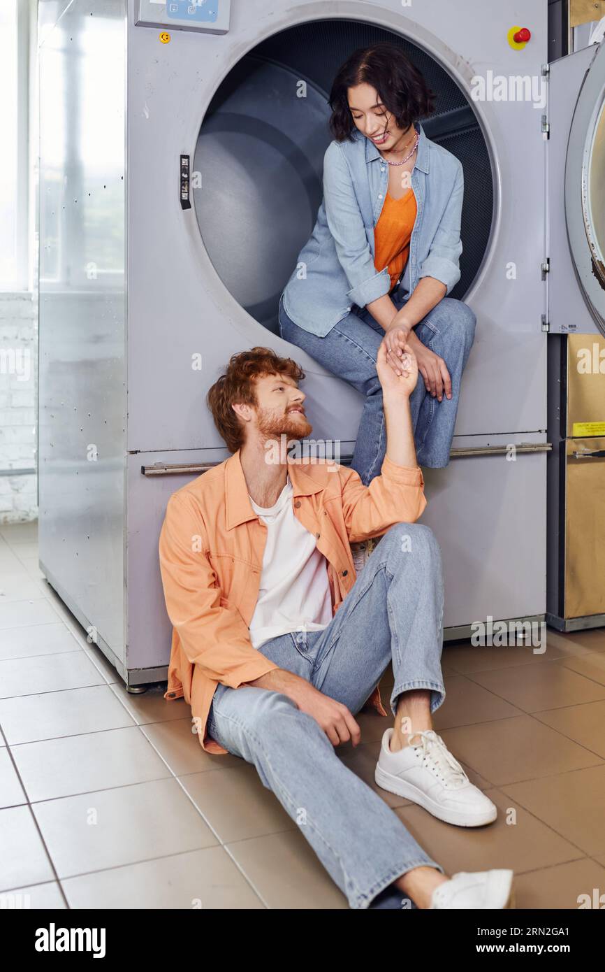 femme asiatique souriante tenant la main de petit ami tout en étant assis sur la machine à laver dans la buanderie à pièces Banque D'Images