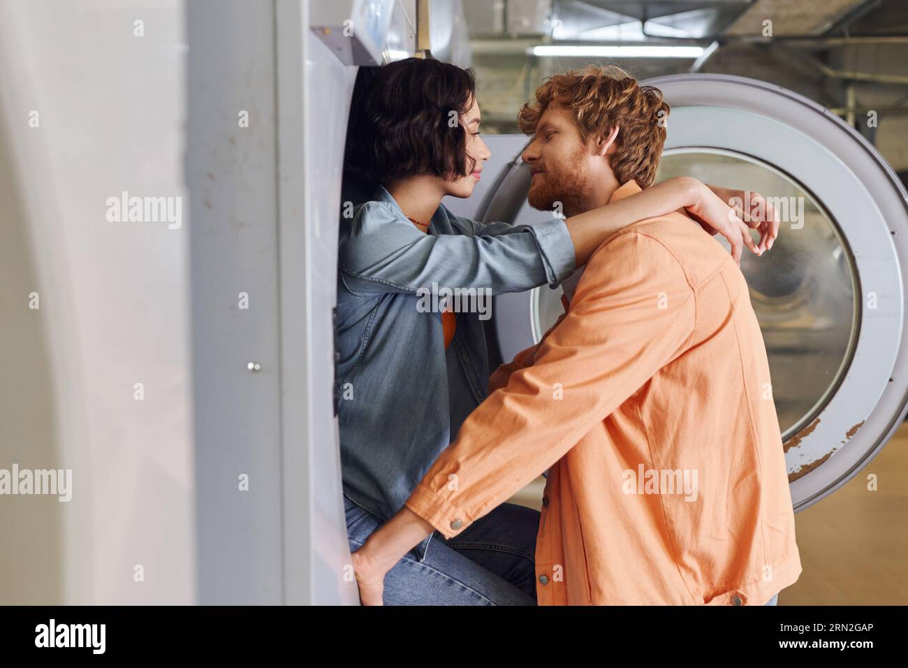 vue latérale de jeune femme asiatique romantique embrassant son petit ami près de la machine à laver dans la buanderie à pièces Banque D'Images