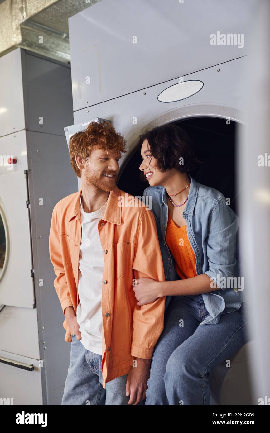 joyeuse jeune femme asiatique embrassant petit ami rousse tout en étant assis sur la machine à laver dans la blanchisserie Banque D'Images