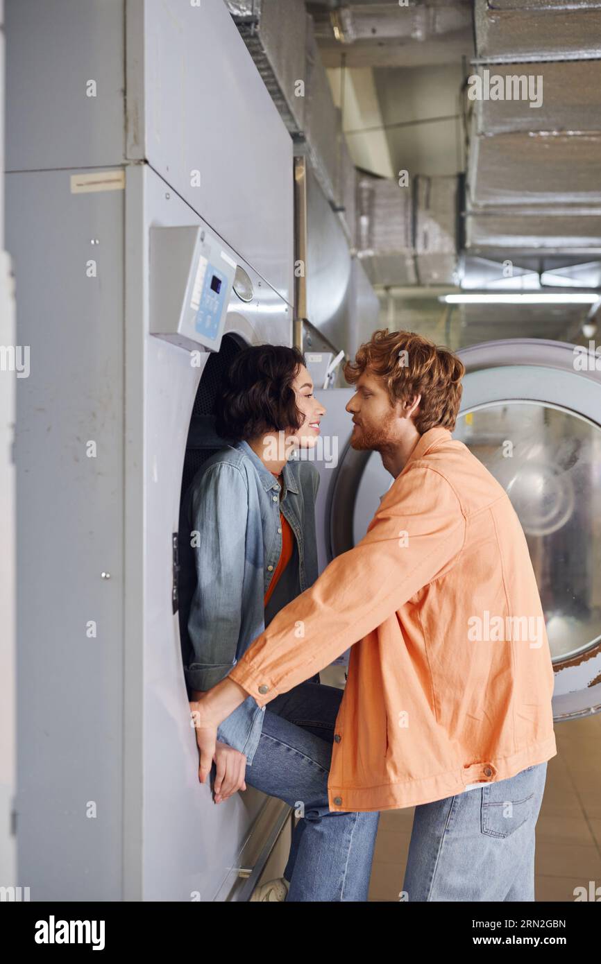 vue latérale de femme asiatique souriante assise sur la machine à laver près de petit ami romantique dans la buanderie à pièces Banque D'Images