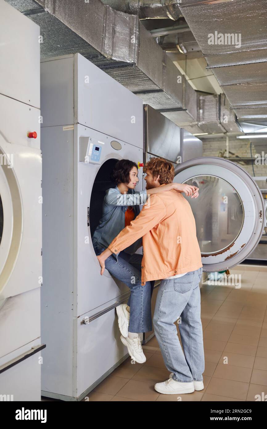 souriant jeune femme asiatique embrassant son petit ami tout en étant assis sur la machine à laver dans la blanchisserie publique Banque D'Images