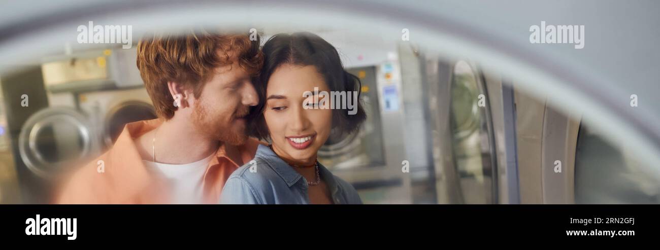 jeune homme rousse chuchotant à une petite amie asiatique souriante dans une blanchisserie publique floue, bannière Banque D'Images