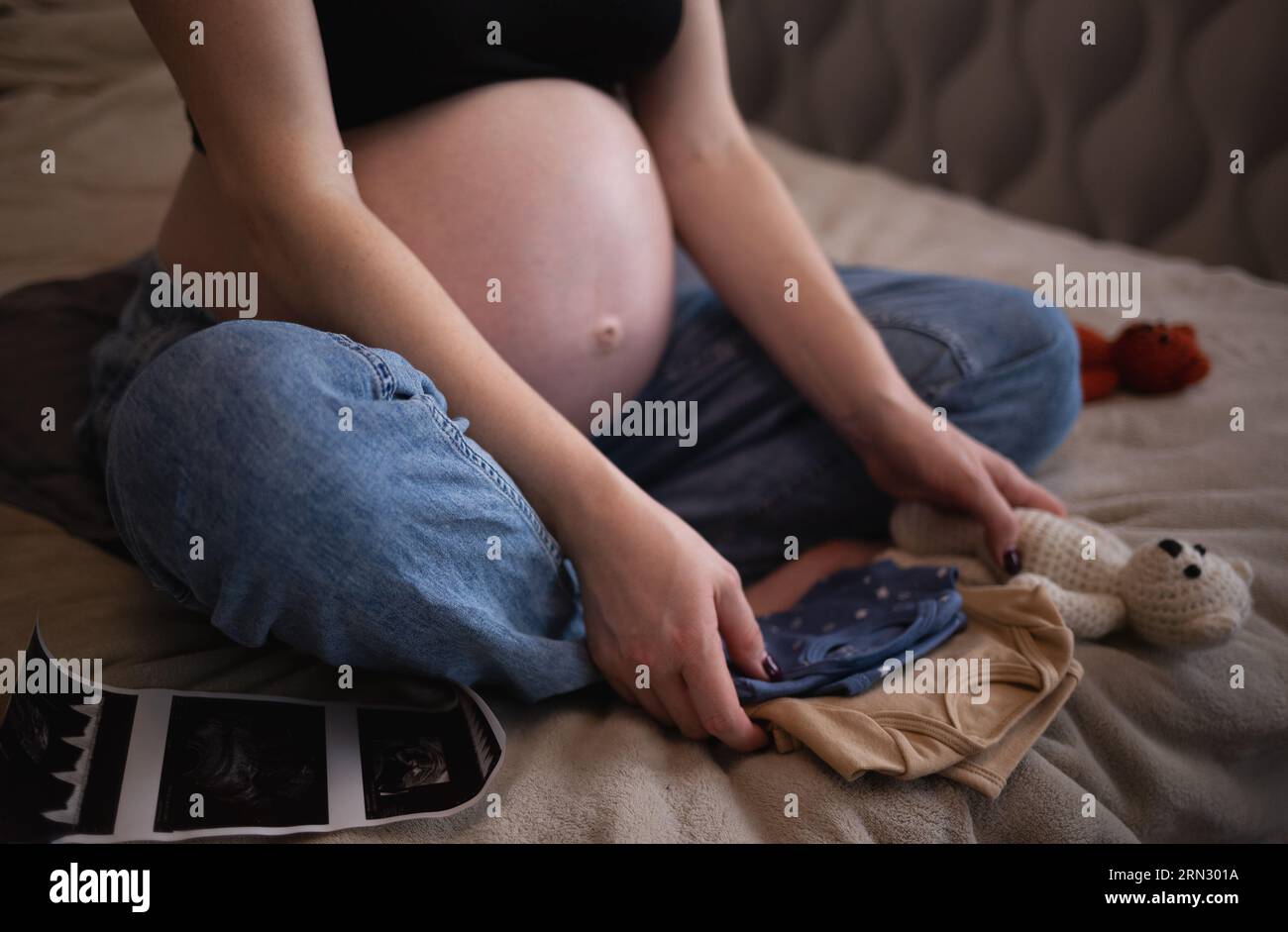 belle jeune femme enceinte est assise sur le lit dans la chambre et tient les petits vêtements de son bébé dans ses mains Banque D'Images