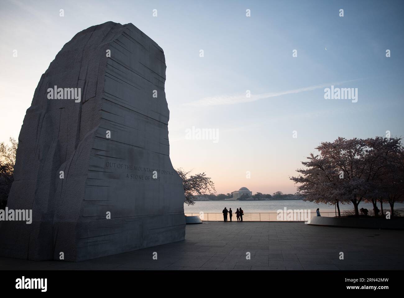 WASHINGTON DC, États-Unis — Un petit groupe de visiteurs se dresse au bord du Tidal Basin avant le lever du soleil, éclipsé par l'imposante sculpture Stone of Hope au Martin Luther King Jr. Memorial. Le Jefferson Memorial est visible au loin à travers les eaux calmes, créant une scène emblématique de Washington DC dans la lumière d'avant l'aube. Banque D'Images