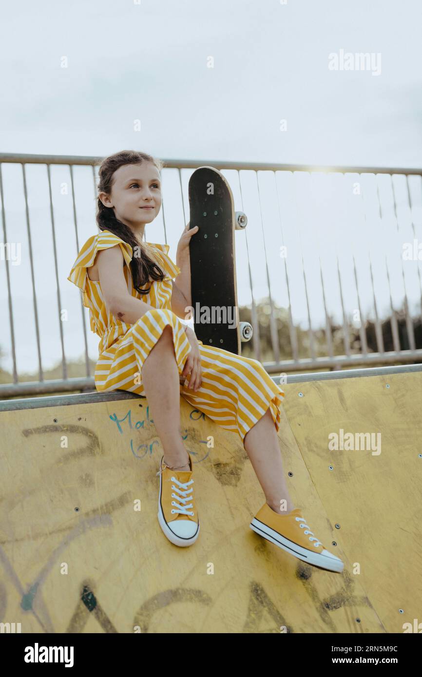 Fille, 9 ans, en combinaison rayée jaune et blanche avec skateboard sur une piste de skateur jaune, mode Banque D'Images