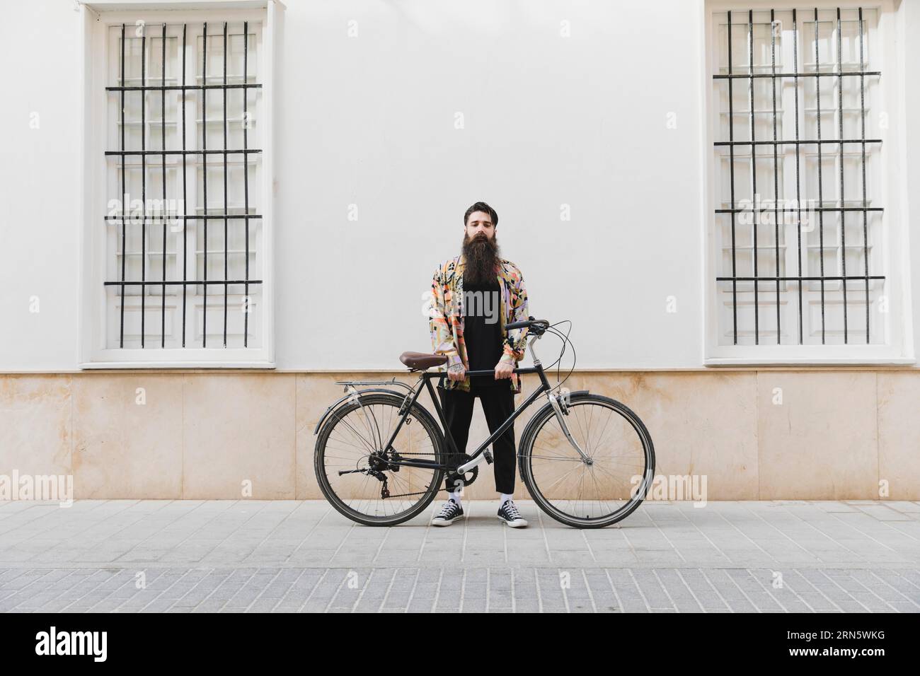 Jeune homme barbu debout avec la bicyclette contre le mur Banque D'Images
