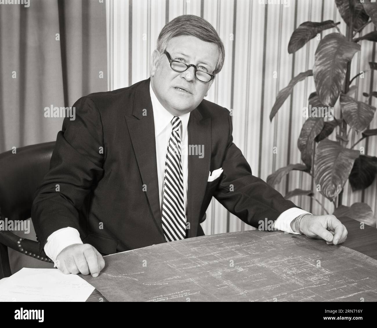 HOMME D'AFFAIRES SÉRIEUX DES ANNÉES 1970 À DESK AVEC DES PLANS PORTANT DES LUNETTES REGARDANT LA CAMÉRA EXPRESSION SÉRIEUSE - O3827 HAR001 HARS B&W MOYEN ÂGE HOMME LIEU DE TRAVAIL CONTACT VISUEL COSTUME COL BLANC ET CRAVATE OCCUPATION DE LEADERSHIP OCCUPATIONS ÉLÉGANT SINCÈRE CRÉATIVITÉ SOLENNELLE CONCENTRÉE PLANTE D'INTÉRIEUR INTENSE NOIR ET BLANC SOIGNEUX ENTREPRENEUR D'ETHNICITÉ CAUCASIENNE EARNEST HAR001 INTENTION DÉMODÉ Banque D'Images