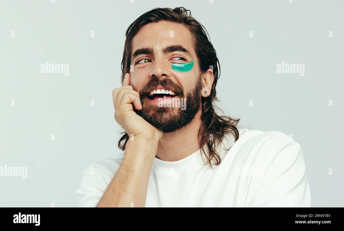 Heureux jeune homme portant un masque sous les yeux dans un studio, dorlotant son visage avec le patch de gel pour réduire les cercles sous les yeux. Beau jeune homme pract Banque D'Images