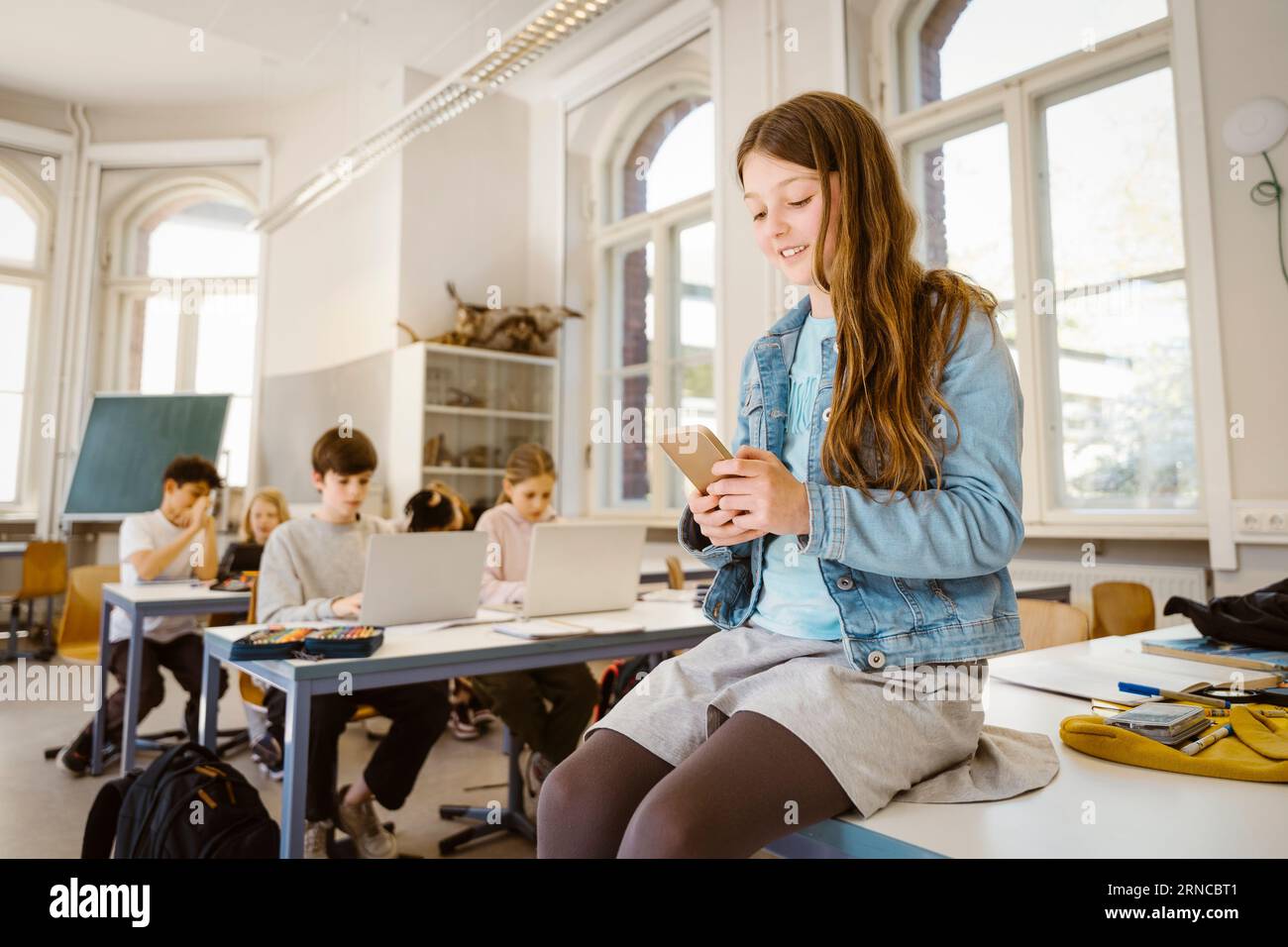 Élève souriant à l'aide d'un téléphone intelligent tout en étant assis au bureau dans la salle de classe Banque D'Images