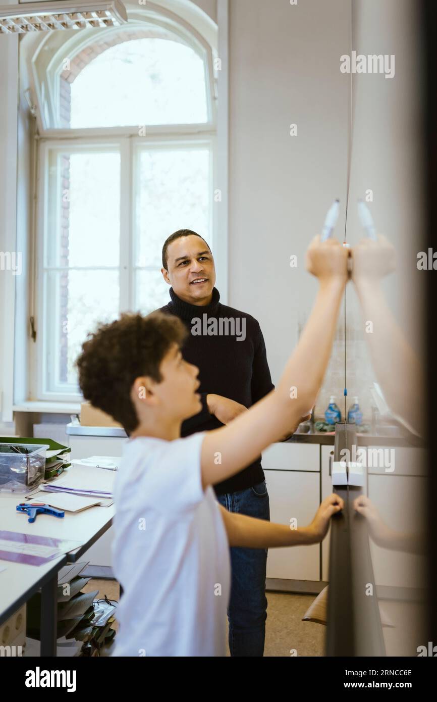 Enseignant masculin regardant l'étudiant écrire sur le tableau blanc dans la salle de classe Banque D'Images