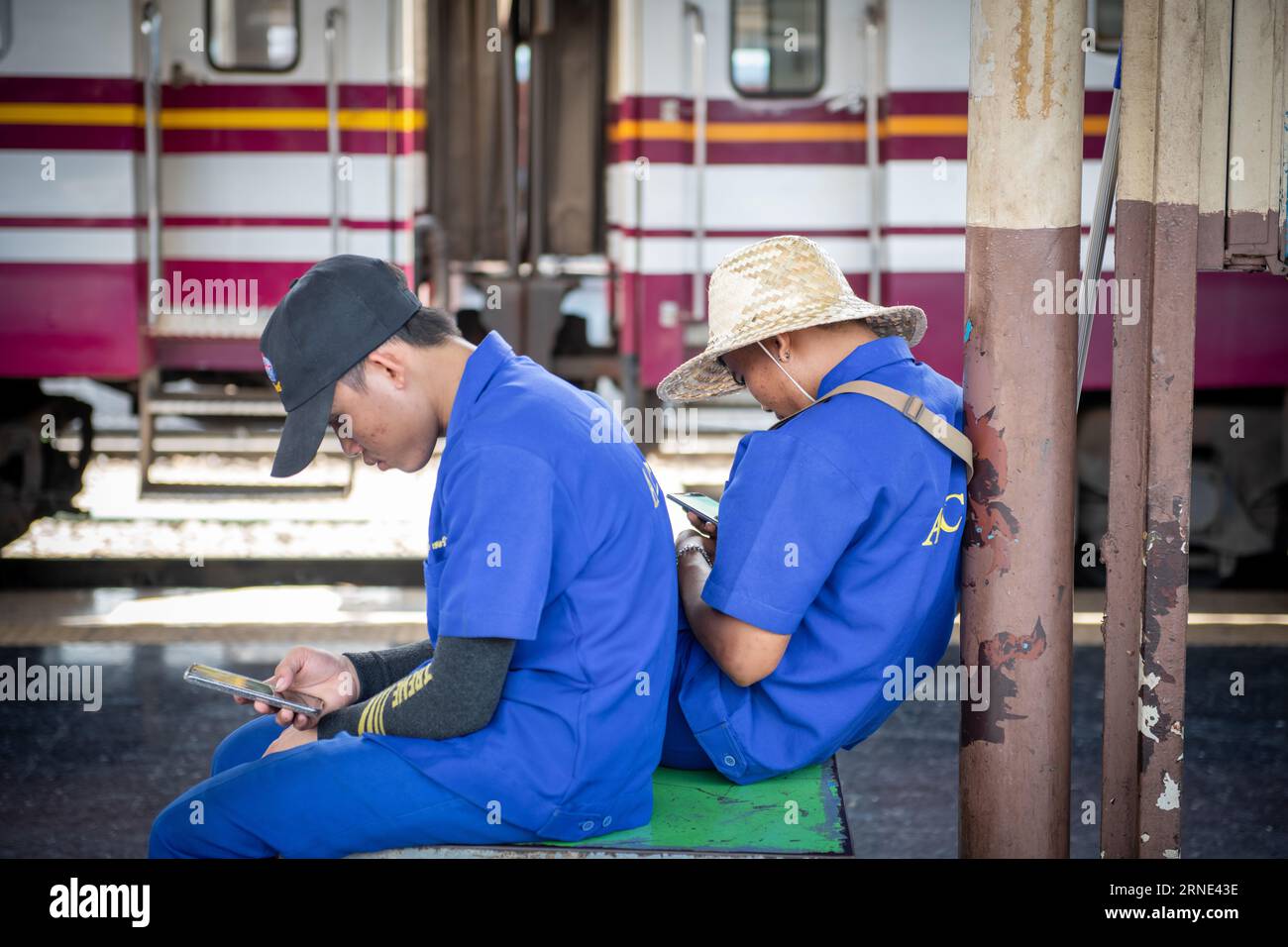 Deux cheminots en uniforme bleu font une pause sur un quai à la gare de Hua Lamphong à Bangkok, en Thaïlande, tout en utilisant leur téléphone portable Banque D'Images
