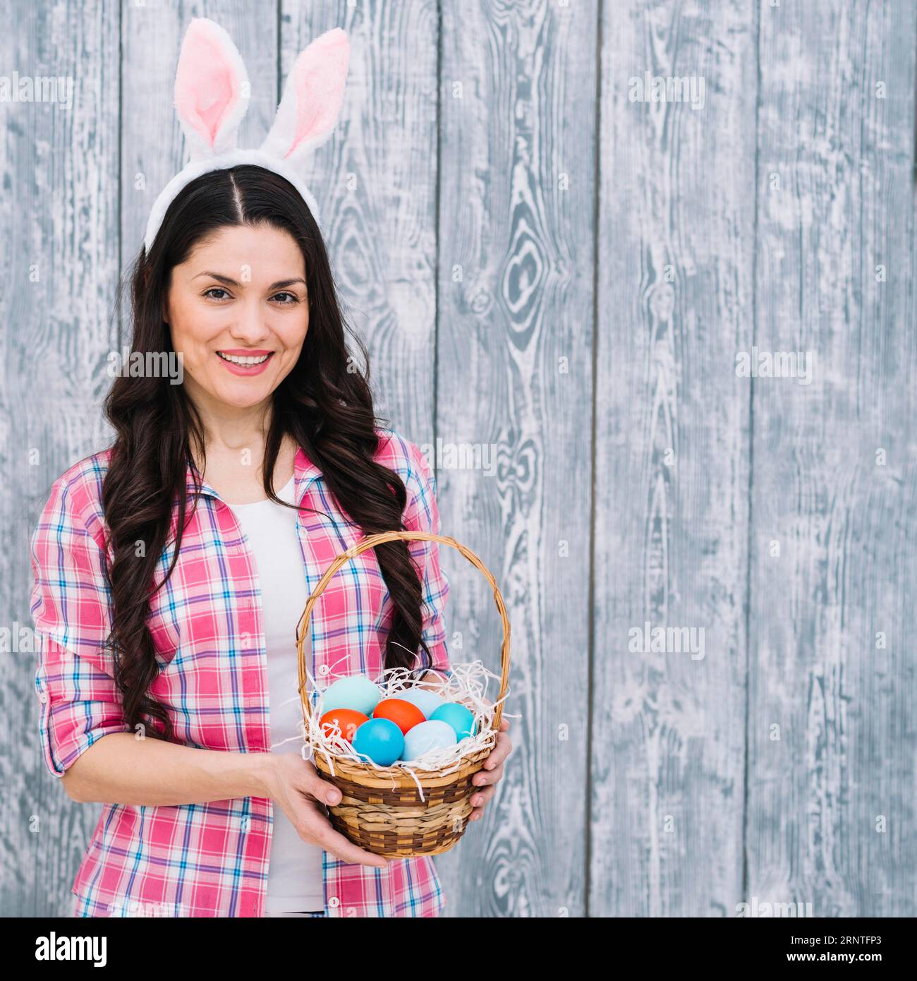 Femme souriante avec tête d'oreilles de lapin montrant le panier d'œufs de pâques sur fond de bois Banque D'Images