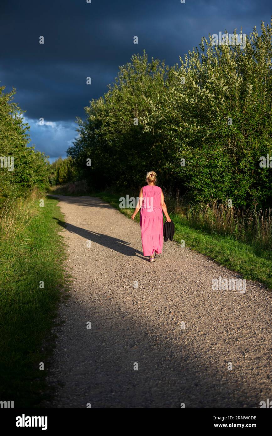 Femme blonde dans une robe rose marchant dans la campagne avec la lumière du soir de longues ombres Banque D'Images